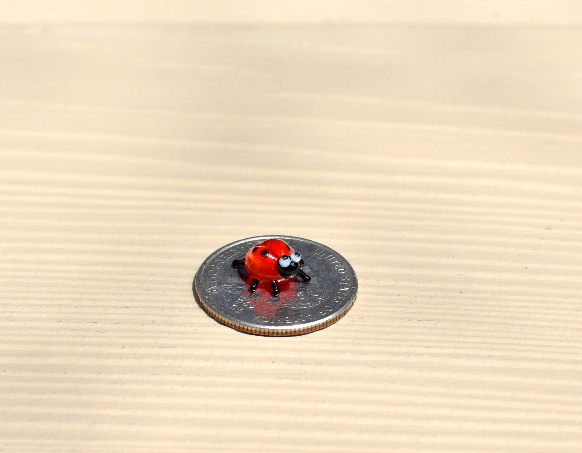 May include: A small red ladybug figurine with black spots and white eyes sitting on a silver US quarter coin.