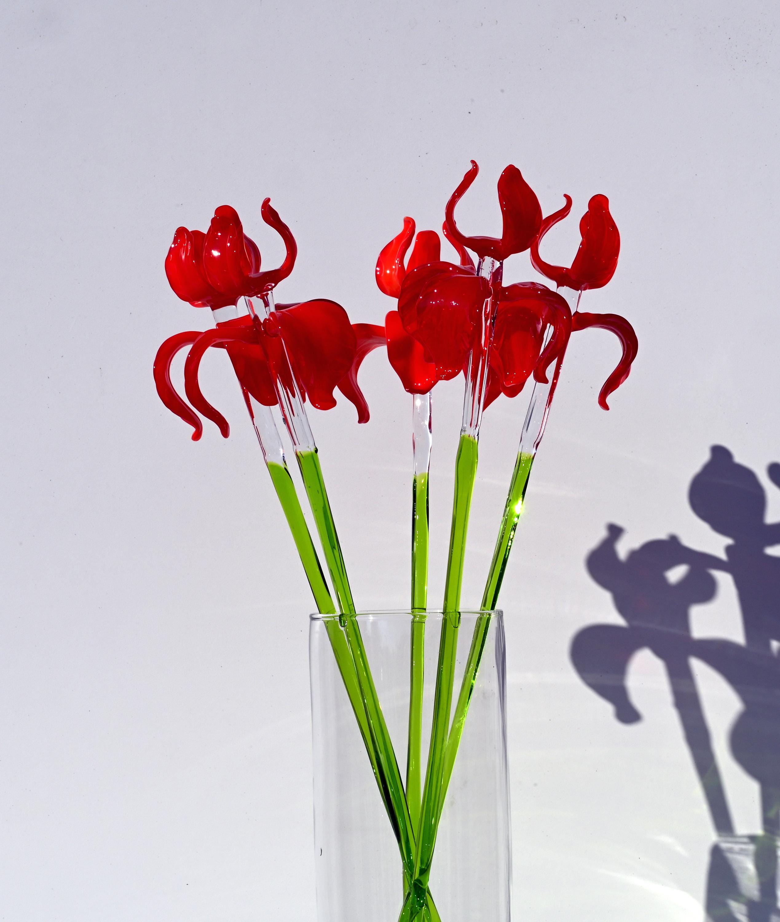 May include: A bouquet of red glass flowers with green stems in a clear glass vase.