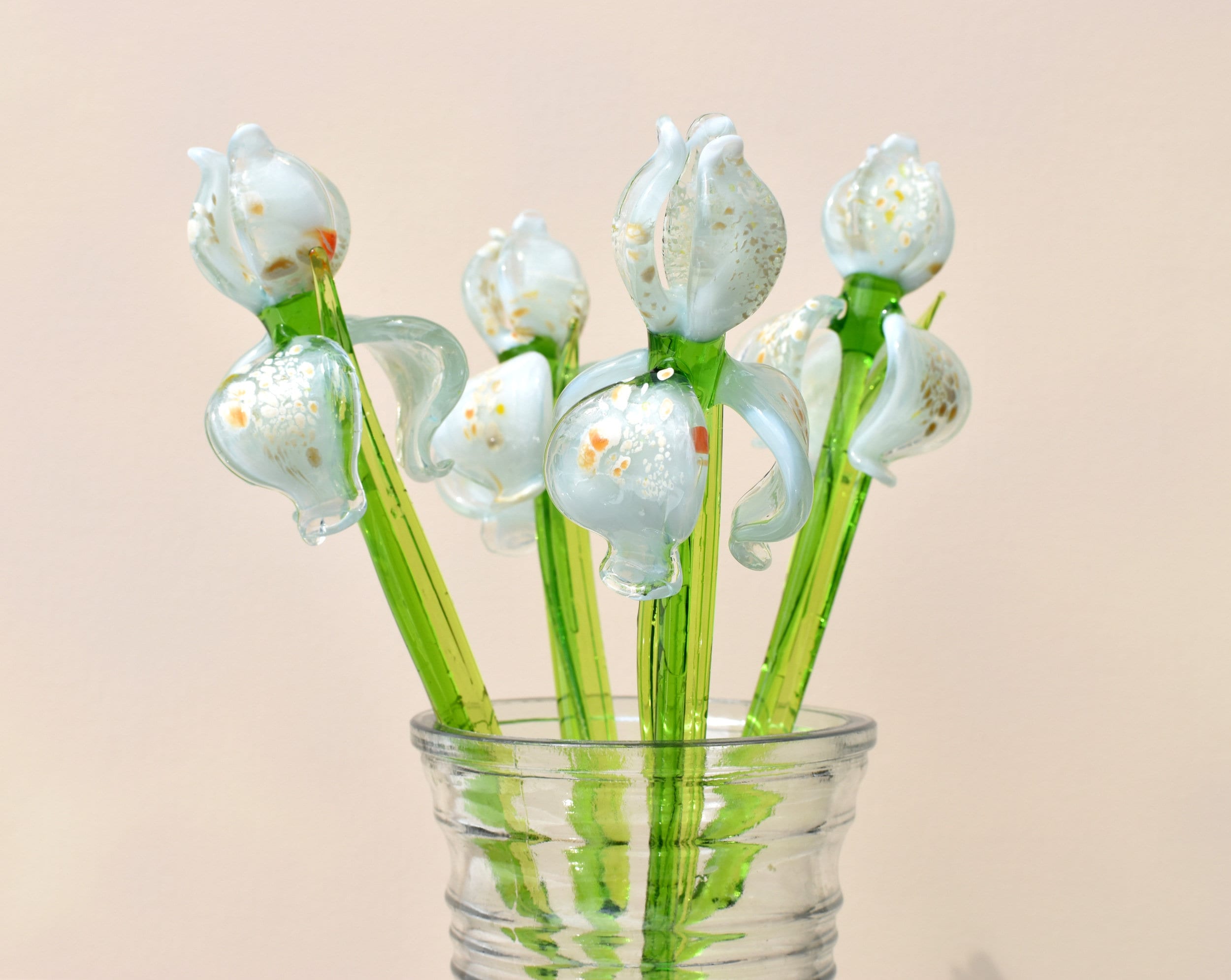 May include: A bouquet of six glass flowers in a clear glass vase. The flowers are white with green stems and have a speckled, iridescent finish. The flowers are arranged in a vase with a textured surface.