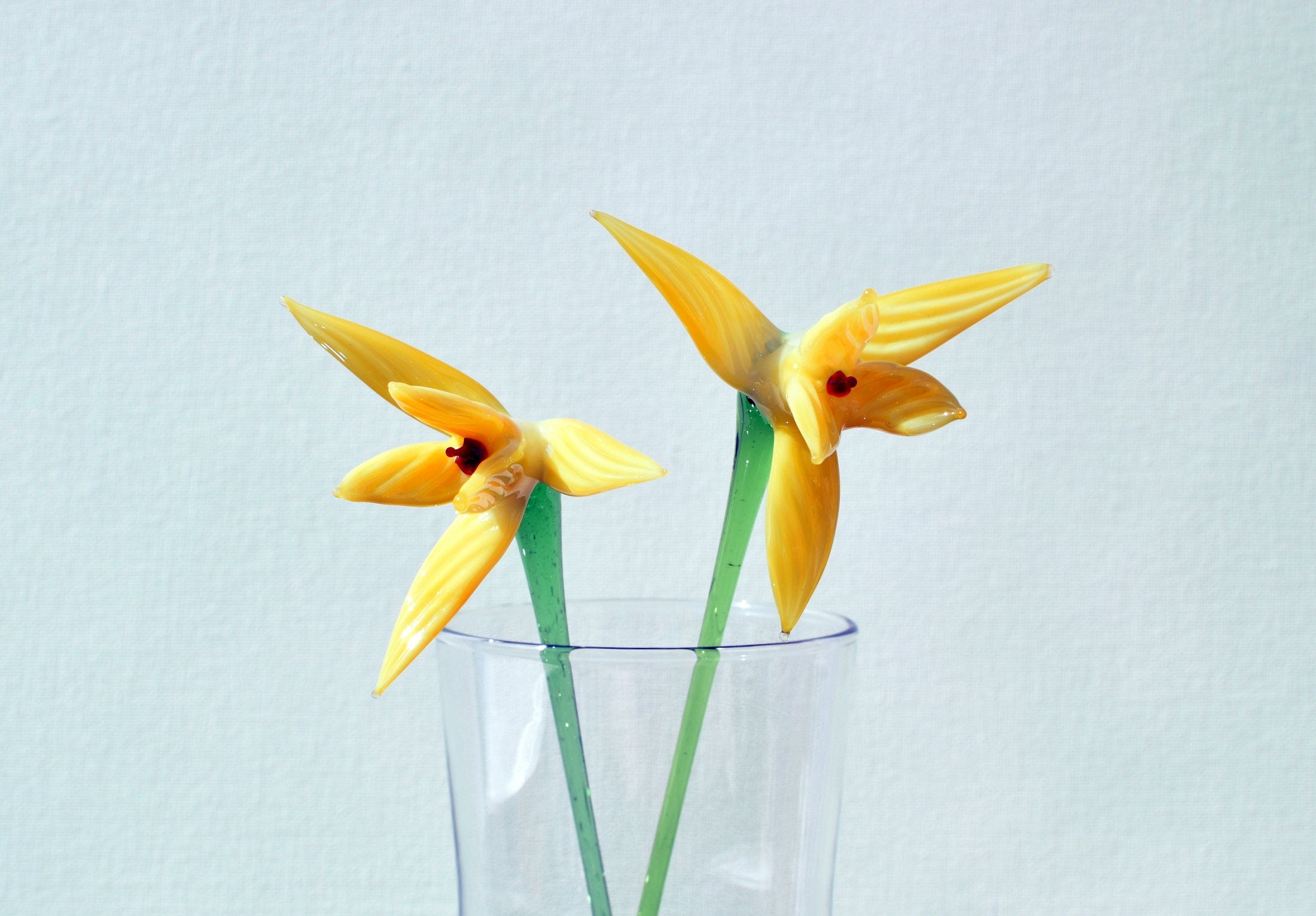 May include: Two yellow glass flowers with green stems in a clear glass vase.