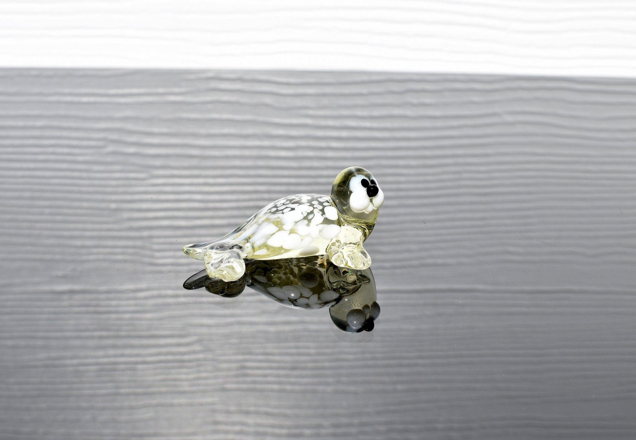 May include: A small glass sea turtle figurine with white and yellow glass. The turtle has black eyes and a white belly. The figurine is sitting on a gray surface.