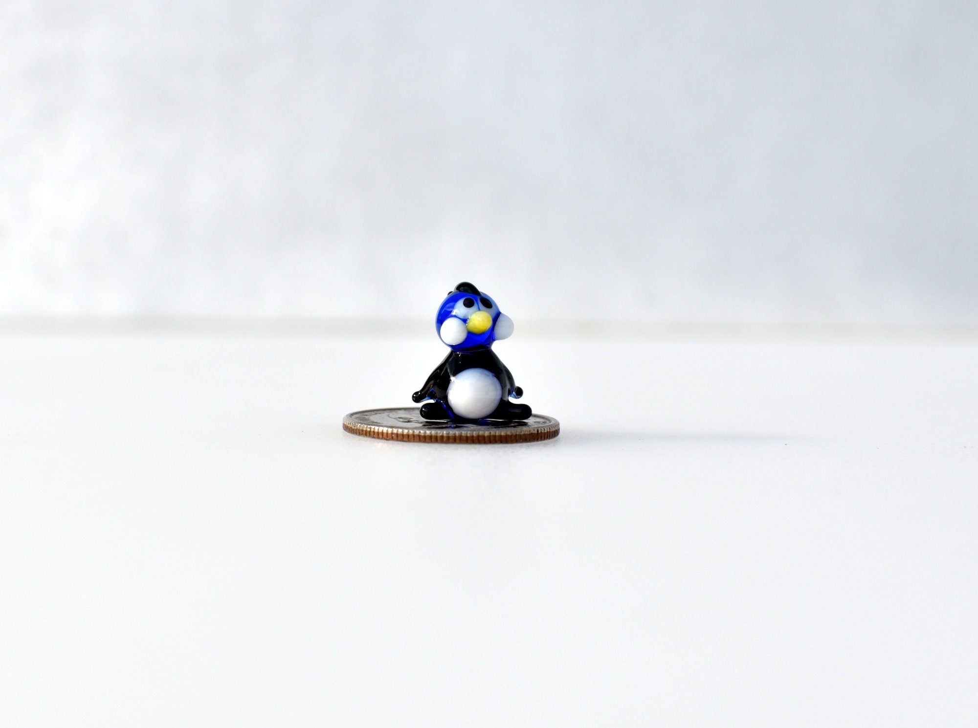 May include: A small blue and white glass penguin figurine sitting on a silver coin.