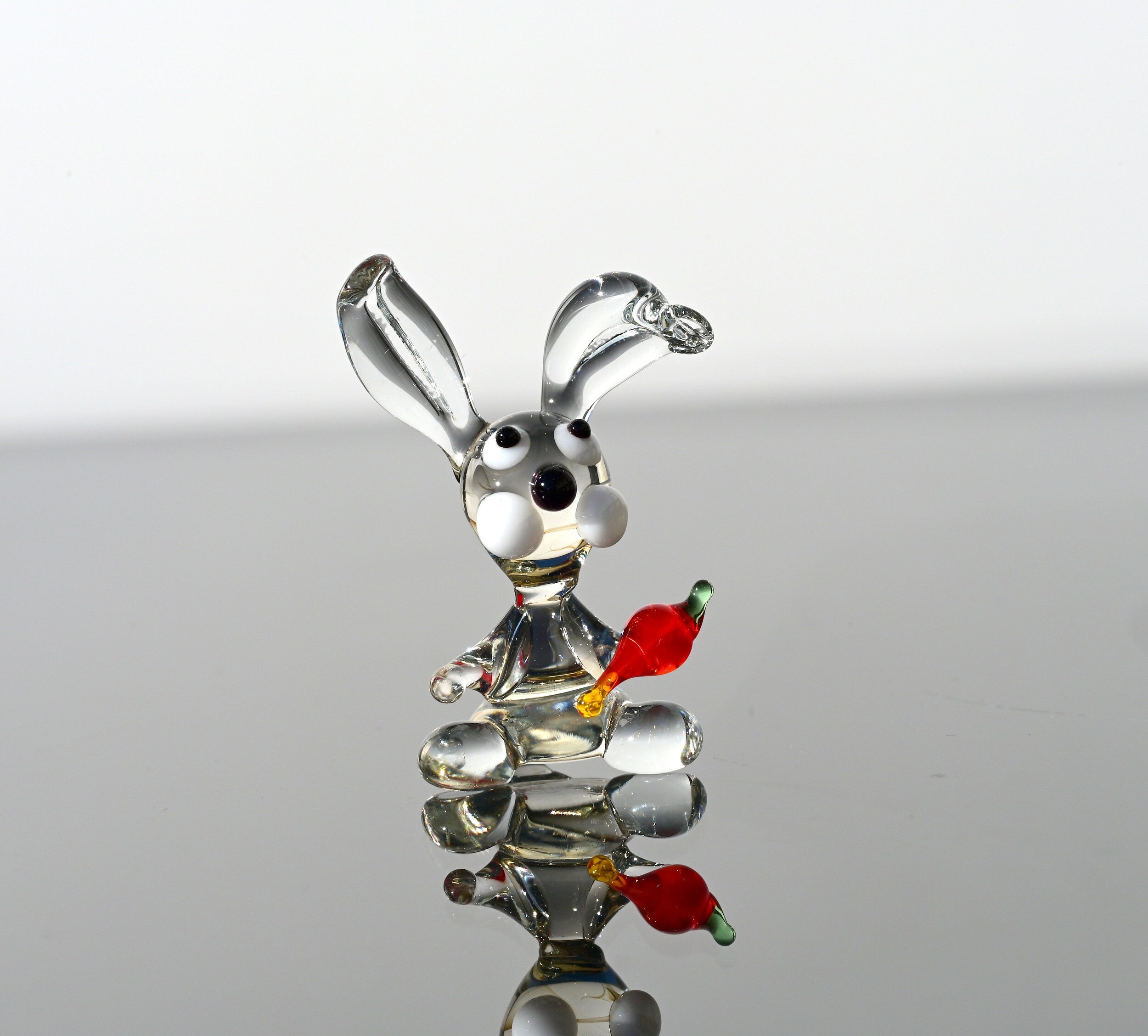 May include: A glass rabbit figurine with a carrot in its mouth. The rabbit is clear glass with black eyes and white accents. The carrot is red and orange glass.