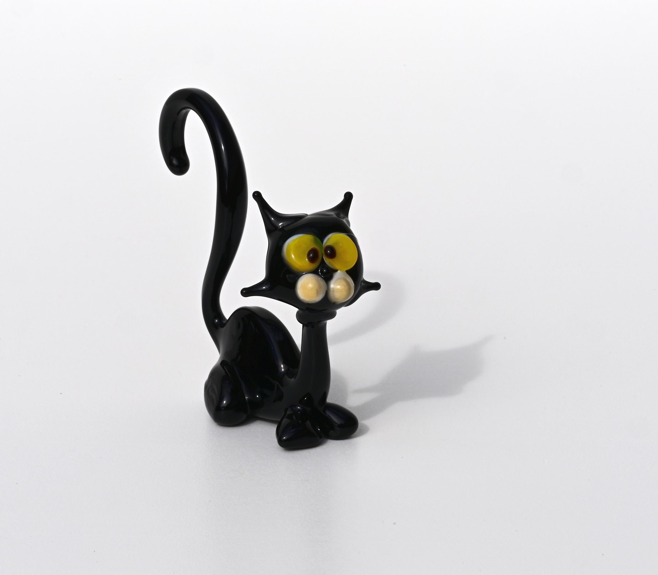 May include: A black glass figurine of a cat with yellow eyes. The cat has a long, curved tail and sits upright with its front legs together. The figurine is a whimsical, cartoonish representation of a black cat, suitable for home decor or as a collectible. The glass is shiny and reflects light, giving it a glossy appearance. The cat's features are exaggerated, with large, expressive eyes and a slightly mischievous expression.