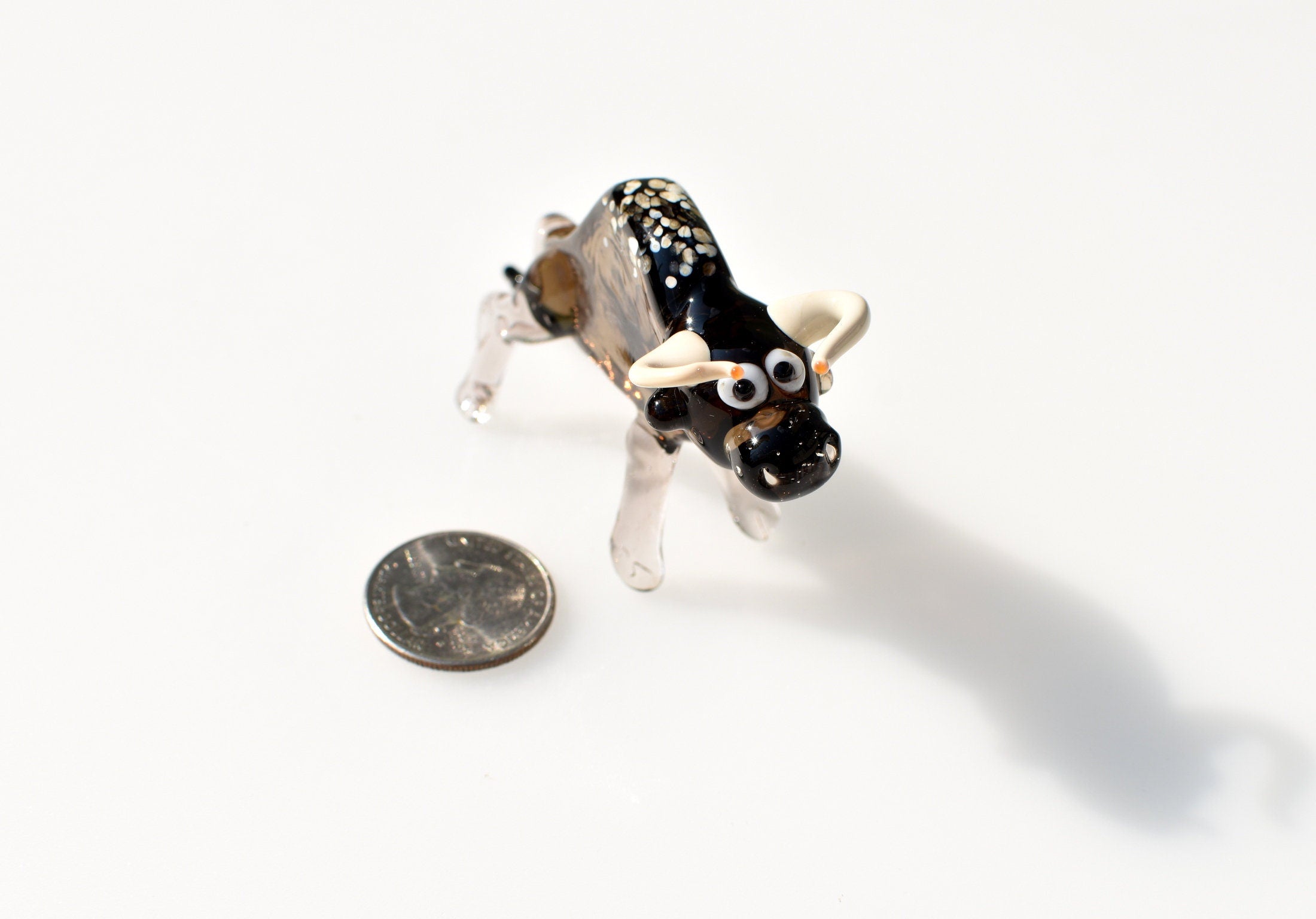 May include: A small glass bull figurine with black and white speckled paint. The bull has large, white eyes with black pupils and small, white horns. The bull is standing on a white surface.