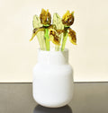 May include: Three glass flowers in a white vase. The flowers are yellow with brown spots and have green stems and leaves.