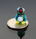 May include: A small glass penguin figurine with a blue and green body, white belly, red beak, and black eyes. The figurine is sitting on a silver US quarter dollar coin.