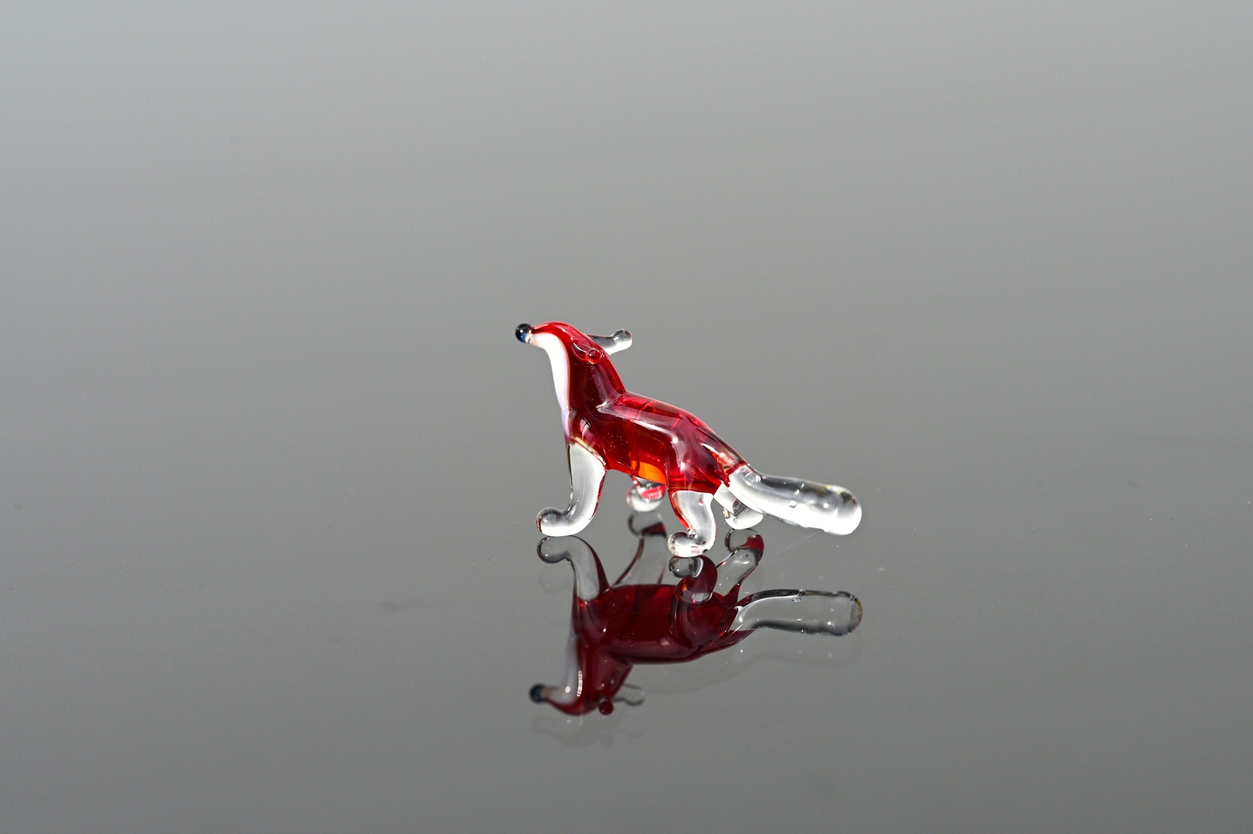May include: A small glass figurine of a fox. The fox is red with a clear glass tail. The figurine is standing on a reflective surface.