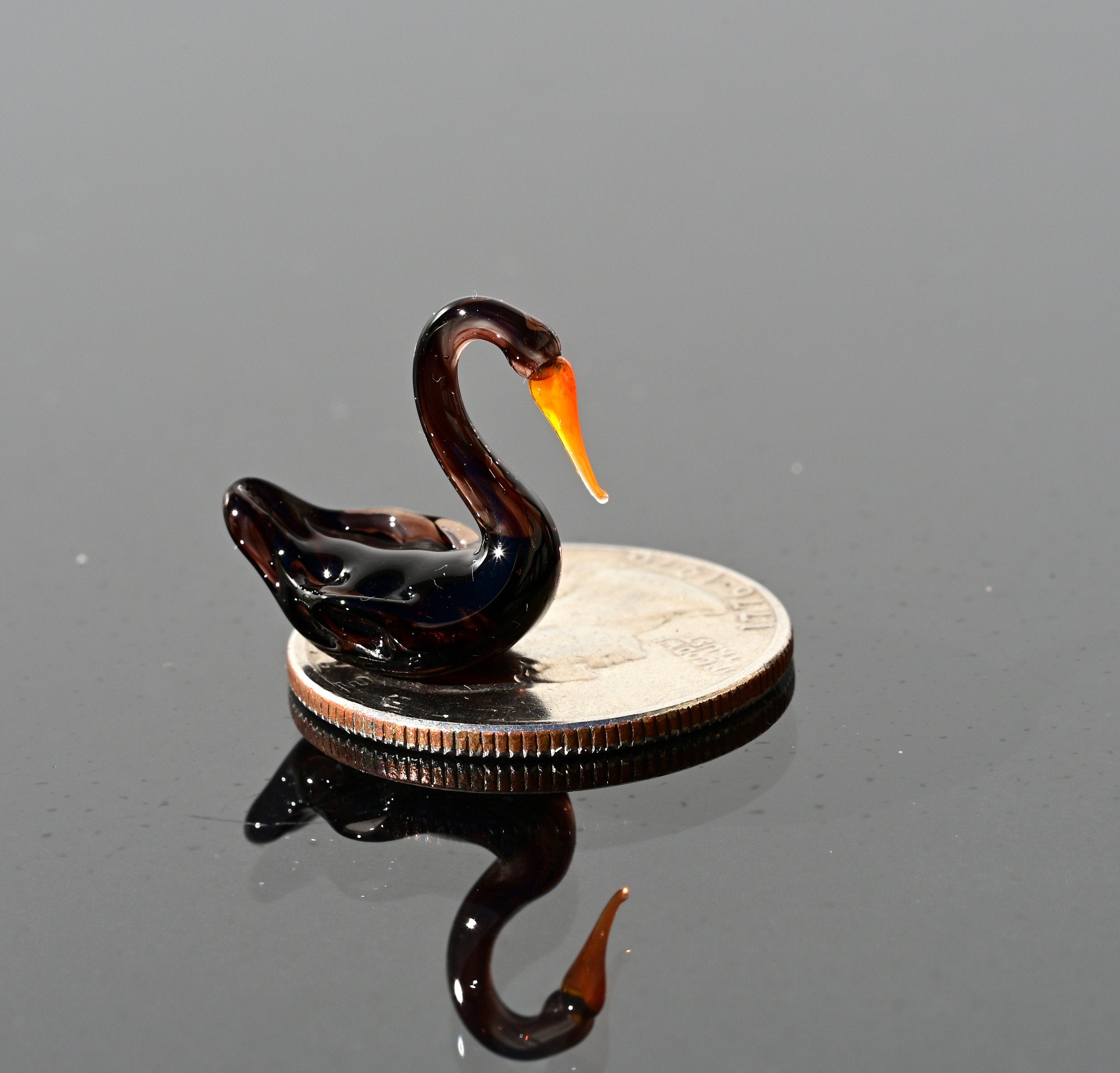 May include: A small black glass swan figurine with an orange beak sitting on a silver US quarter coin.