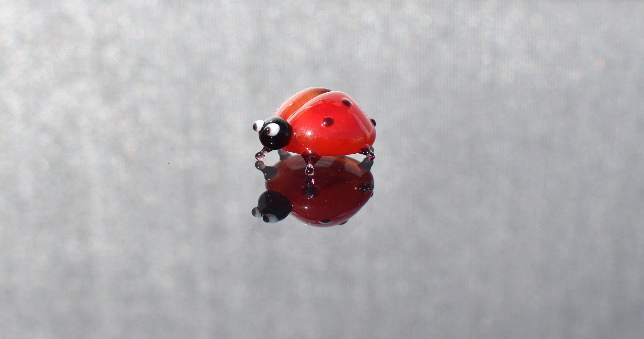 May include: A red and black glass ladybug figurine with white eyes. The ladybug is sitting on a reflective surface.