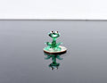 May include: A small green glass frog figurine sitting on a silver coin. The frog has black eyes and is sitting in a sitting position with its legs bent.