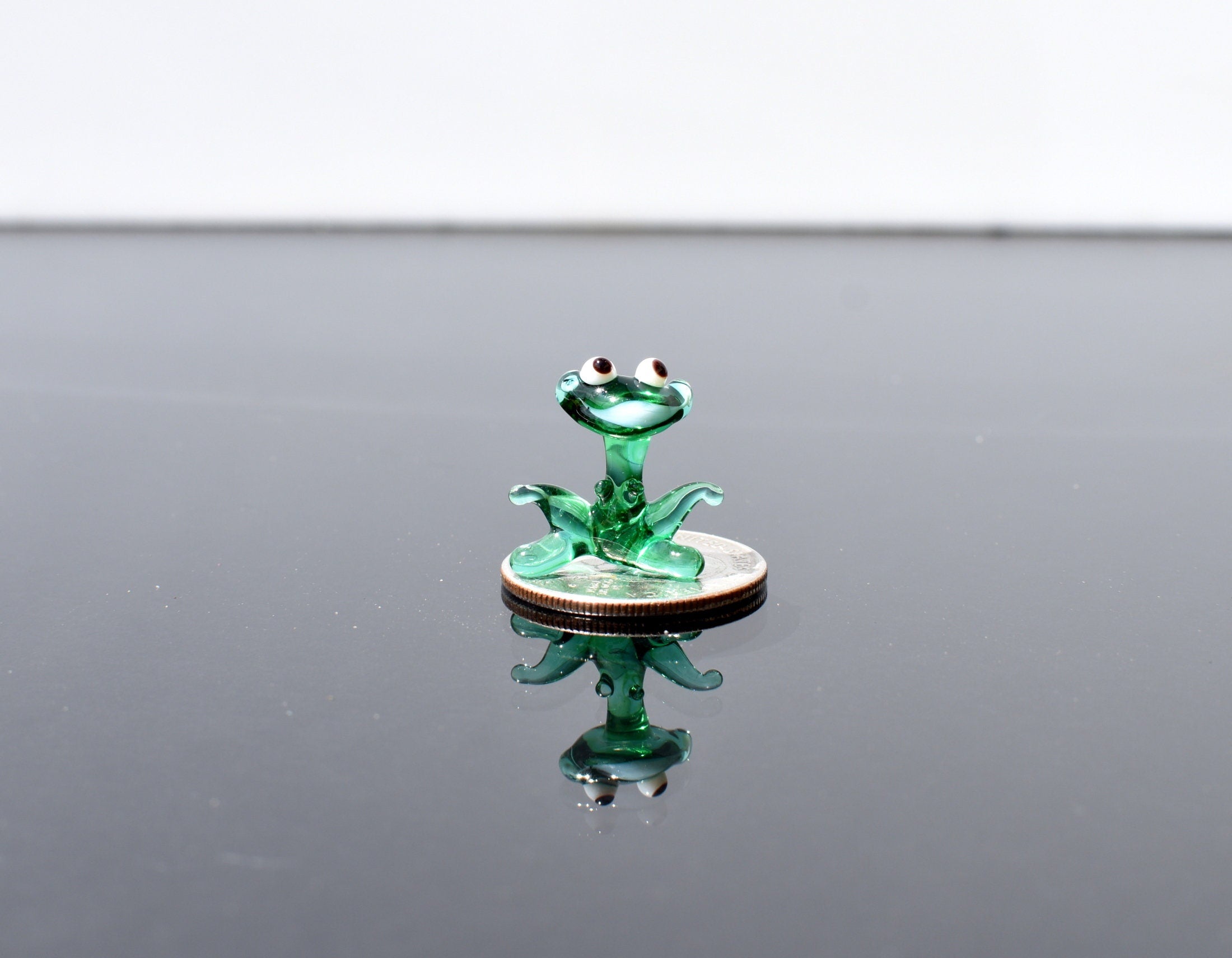 May include: A small green glass frog figurine sitting on a silver coin. The frog has black eyes and is sitting in a sitting position with its legs bent.