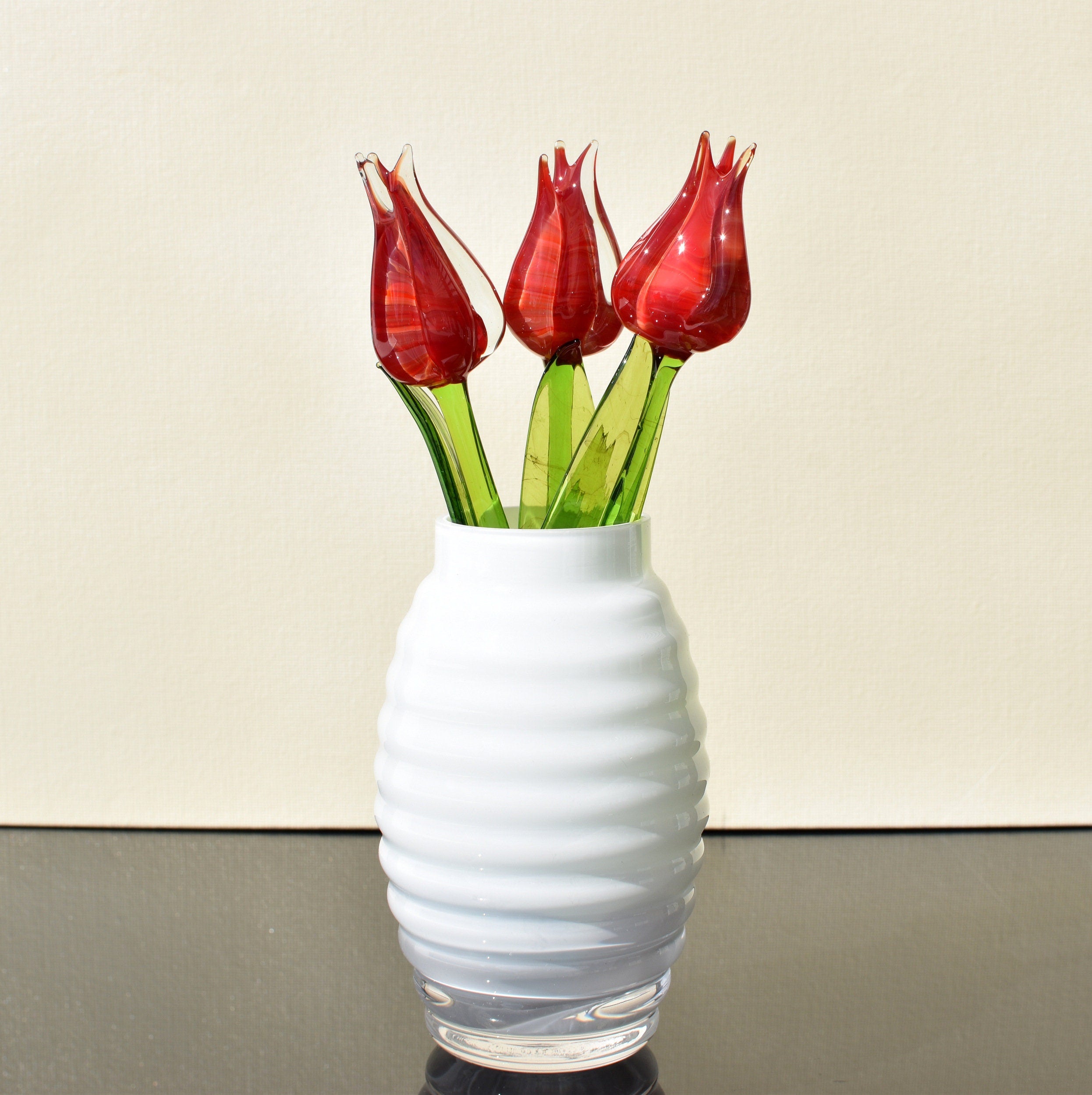 May include: Four red glass tulips with green stems in a white ribbed vase.