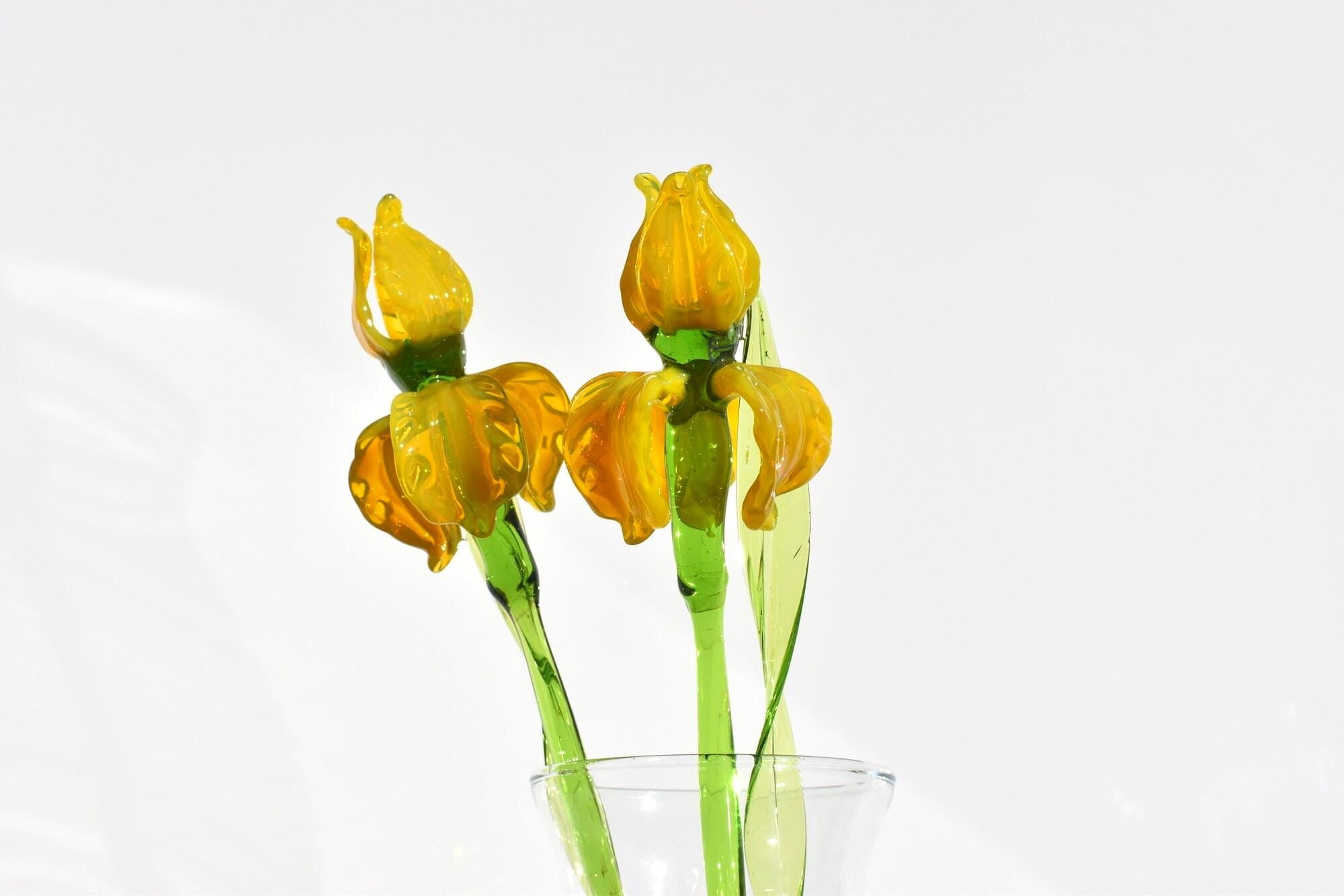 May include: Two yellow glass flowers with green stems and leaves in a clear glass vase.