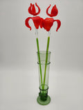 May include: A tall, green glass vase holds two artificial red flowers. The flowers have curved petals and clear stems with green accents. The vase is cylindrical with decorative bands and a circular base, all in a matching green hue. The background is a plain white.