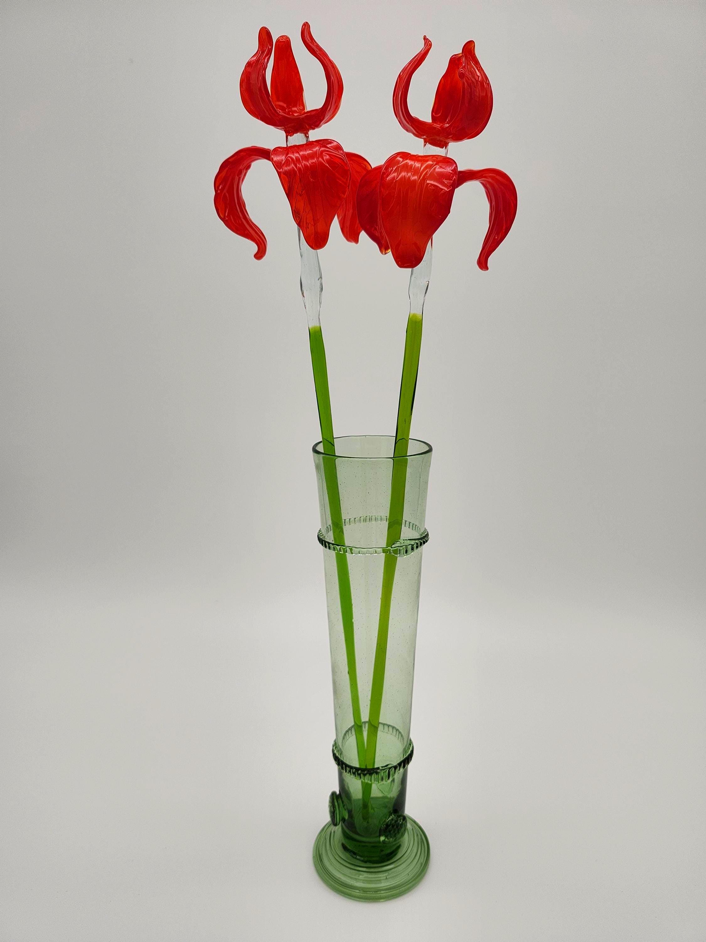 May include: A tall, green glass vase holds two artificial red flowers. The flowers have curved petals and clear stems with green accents. The vase is cylindrical with decorative bands and a circular base, all in a matching green hue. The background is a plain white.