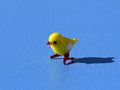 May include: A small yellow glass figurine of a chick with red legs and a white spot on its chest. The chick is standing on a blue surface.