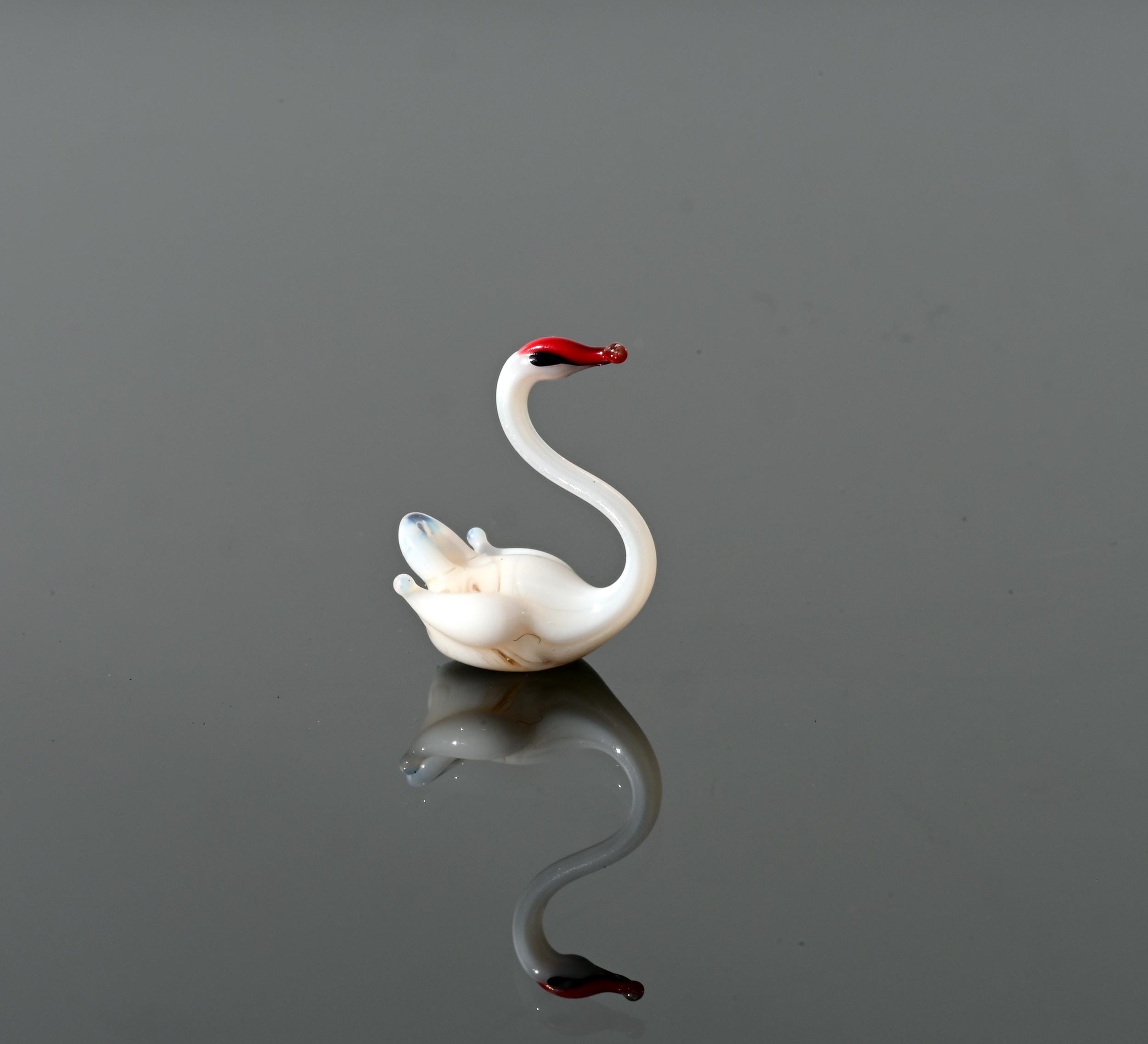 May include: A small white glass swan figurine with a red beak. The swan is in a swimming pose with its head turned to the side.