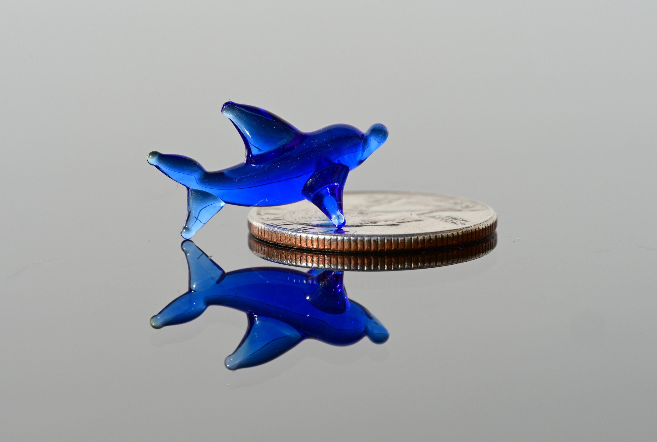 May include: A small blue glass dolphin figurine is positioned on top of a silver US quarter. The dolphin is facing the right side of the image and its reflection is visible on the surface below.