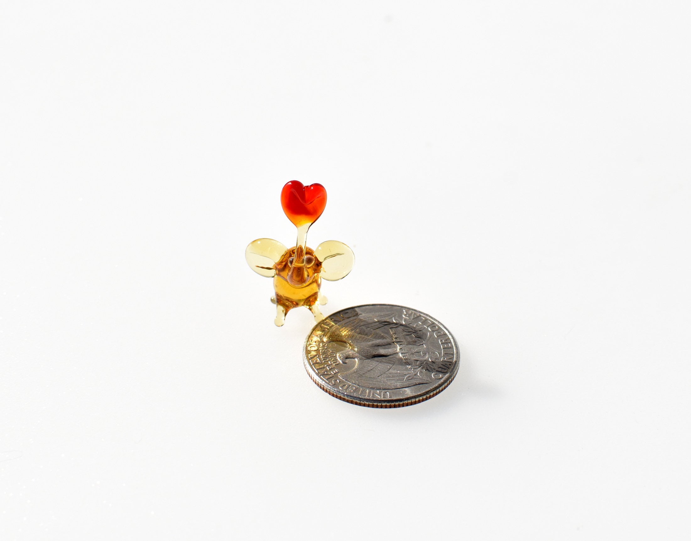 May include: A small glass figurine of a mouse with a red heart on top. The mouse is amber colored and has wings. The figurine is next to a US quarter for size comparison.