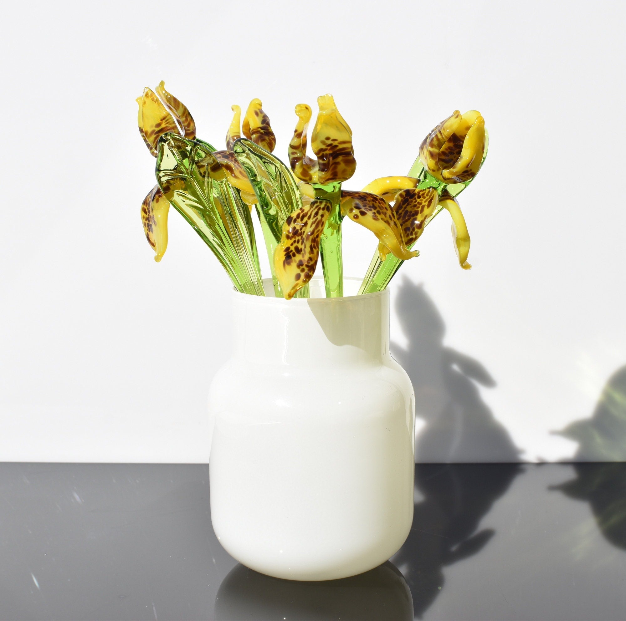 May include: A white ceramic vase with a bouquet of glass flowers. The flowers are yellow with brown spots and green stems.