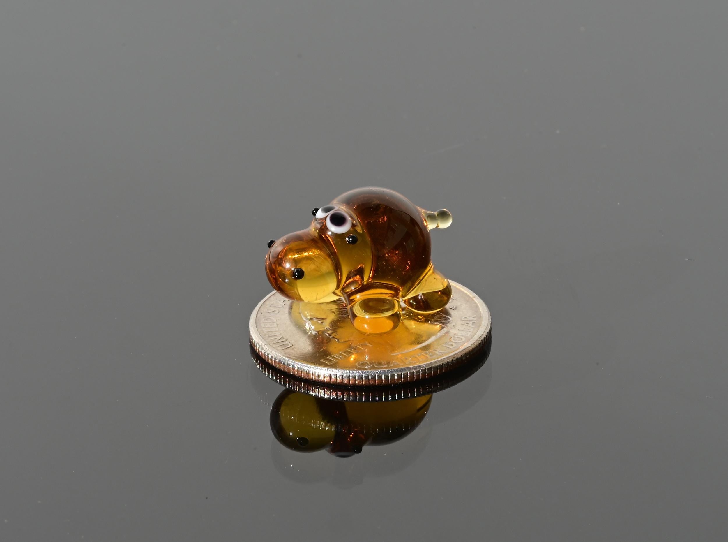 May include: A small amber glass figurine of a dog sitting on a silver coin. The dog has black eyes and a brown body.