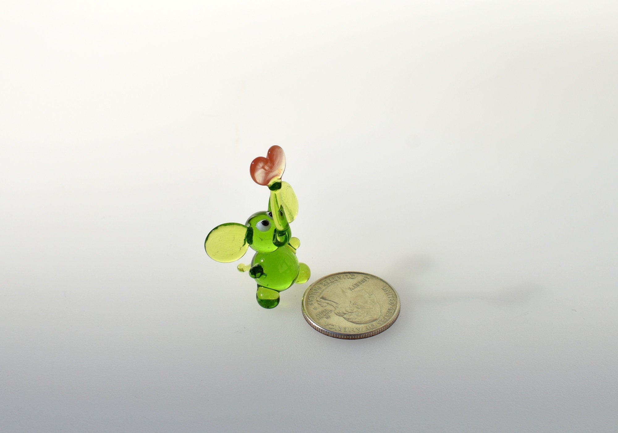 May include: A small green glass elephant figurine holding a red heart. The elephant is standing on a white surface next to a US quarter.