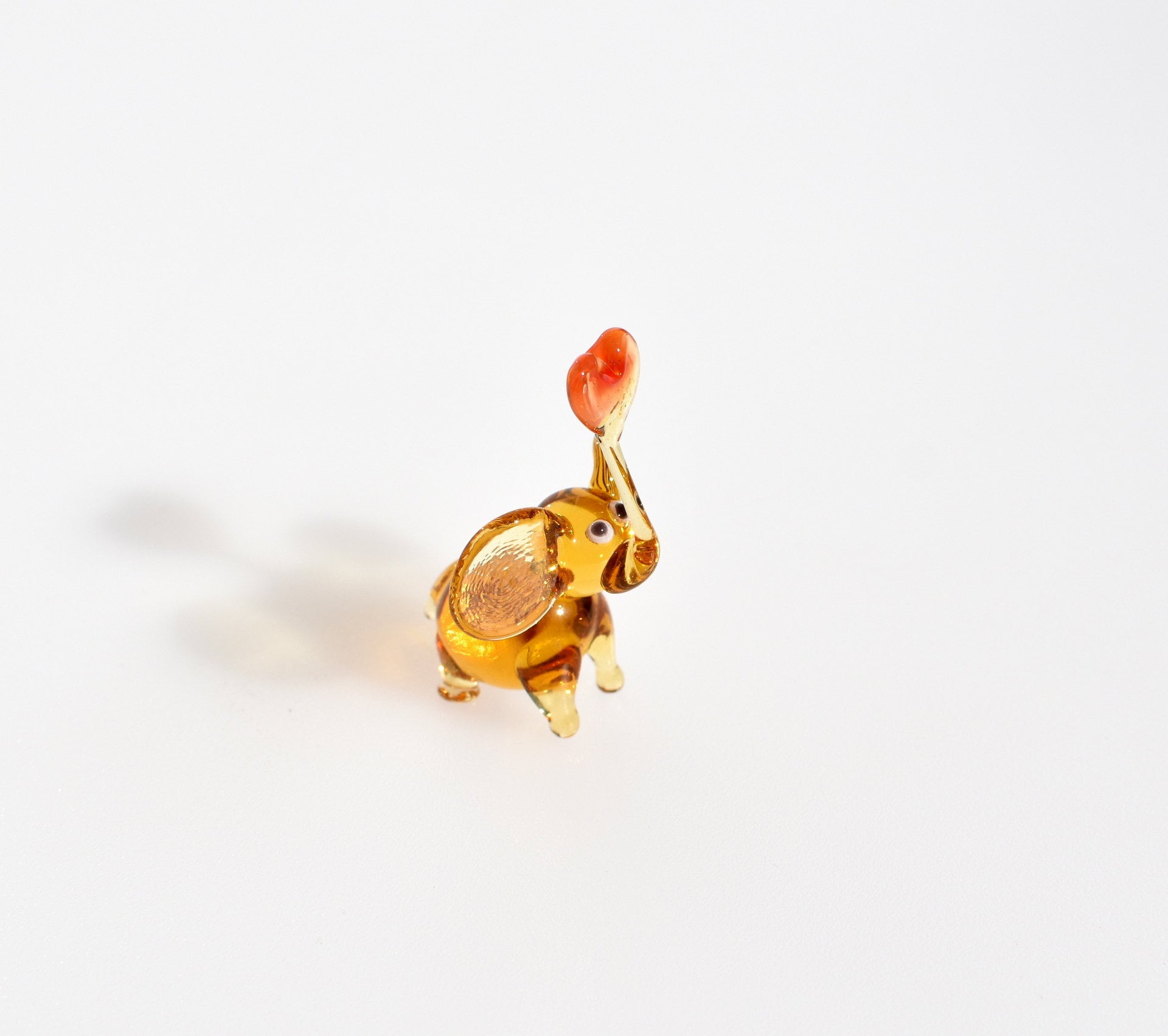 May include: A small glass elephant figurine with a heart-shaped trunk. The elephant is amber in color with white eyes and a red heart on the top of its trunk.