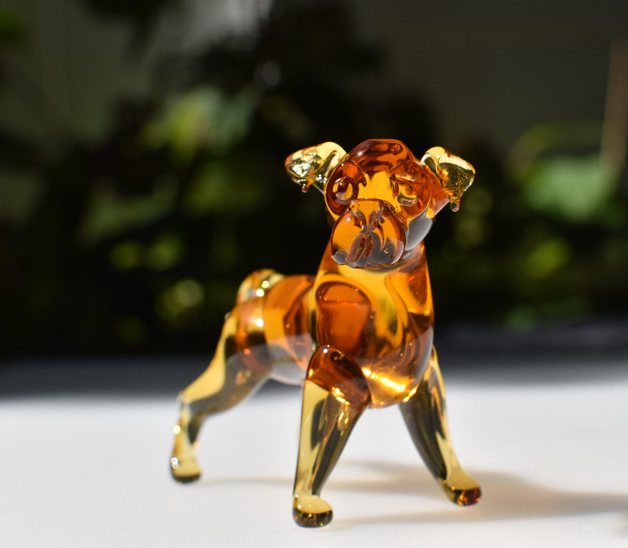 May include: A glass figurine of a pug dog. The figurine is amber colored and has a detailed face and body. The figurine is standing on a white surface.
