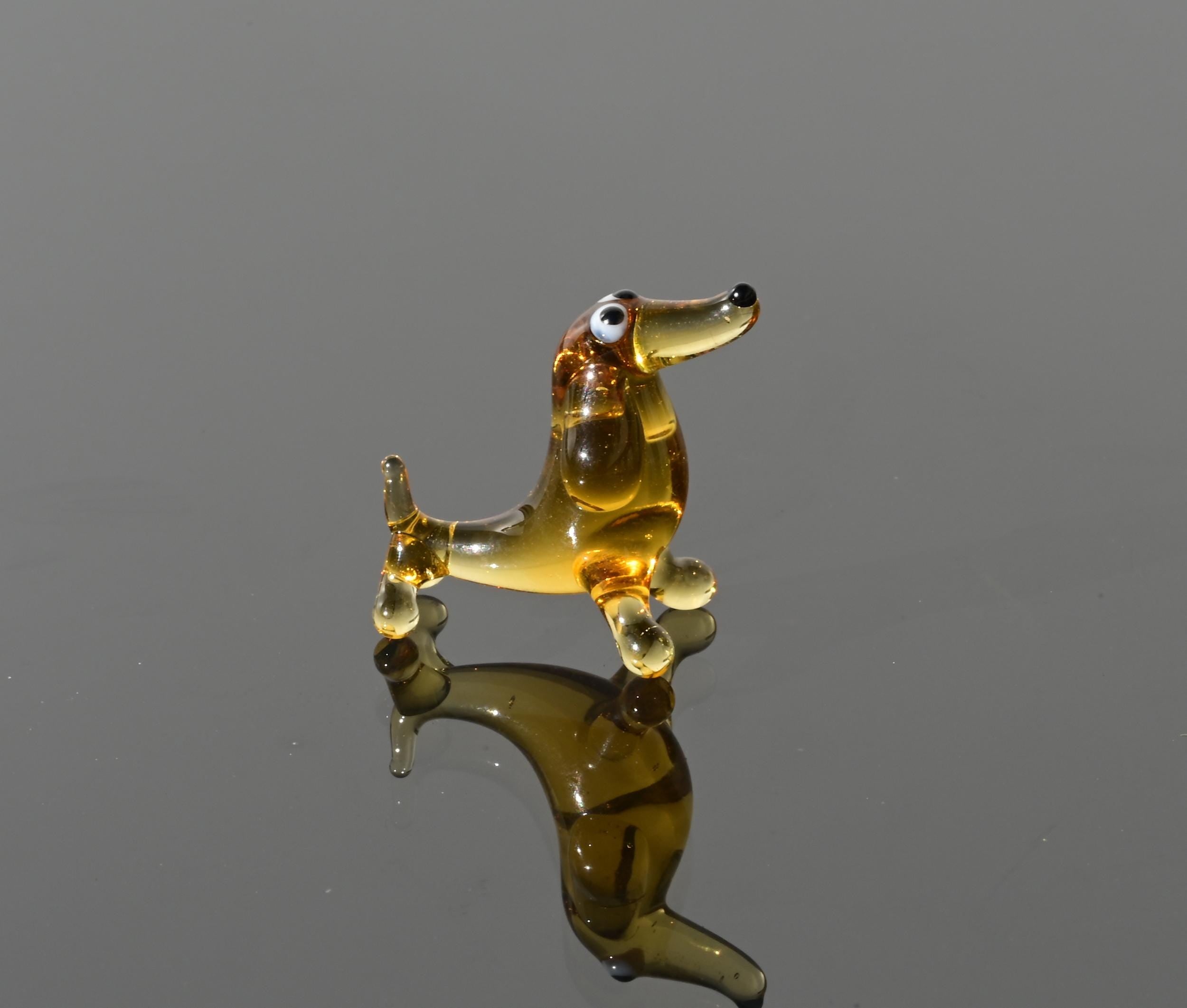 May include: A small glass dachshund figurine in amber color with black eyes and a black nose. The figurine is standing on a reflective surface.