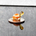 May include: A small glass turtle figurine with a yellow head and orange and white shell. The turtle is sitting on a silver coin.