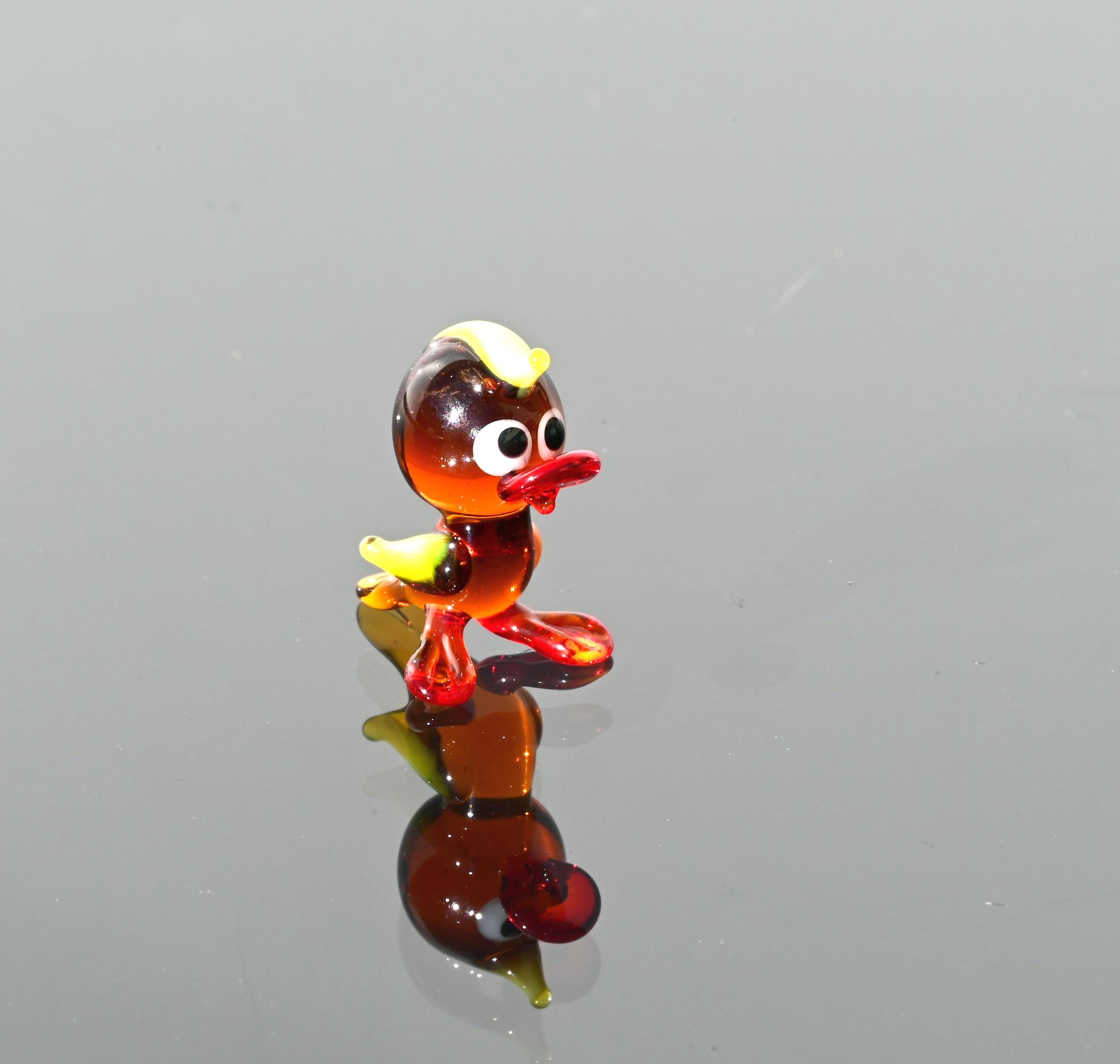 May include: A small glass duck figurine with a yellow beak, red feet, and brown body. The duck has large, black eyes and is standing on a reflective surface.