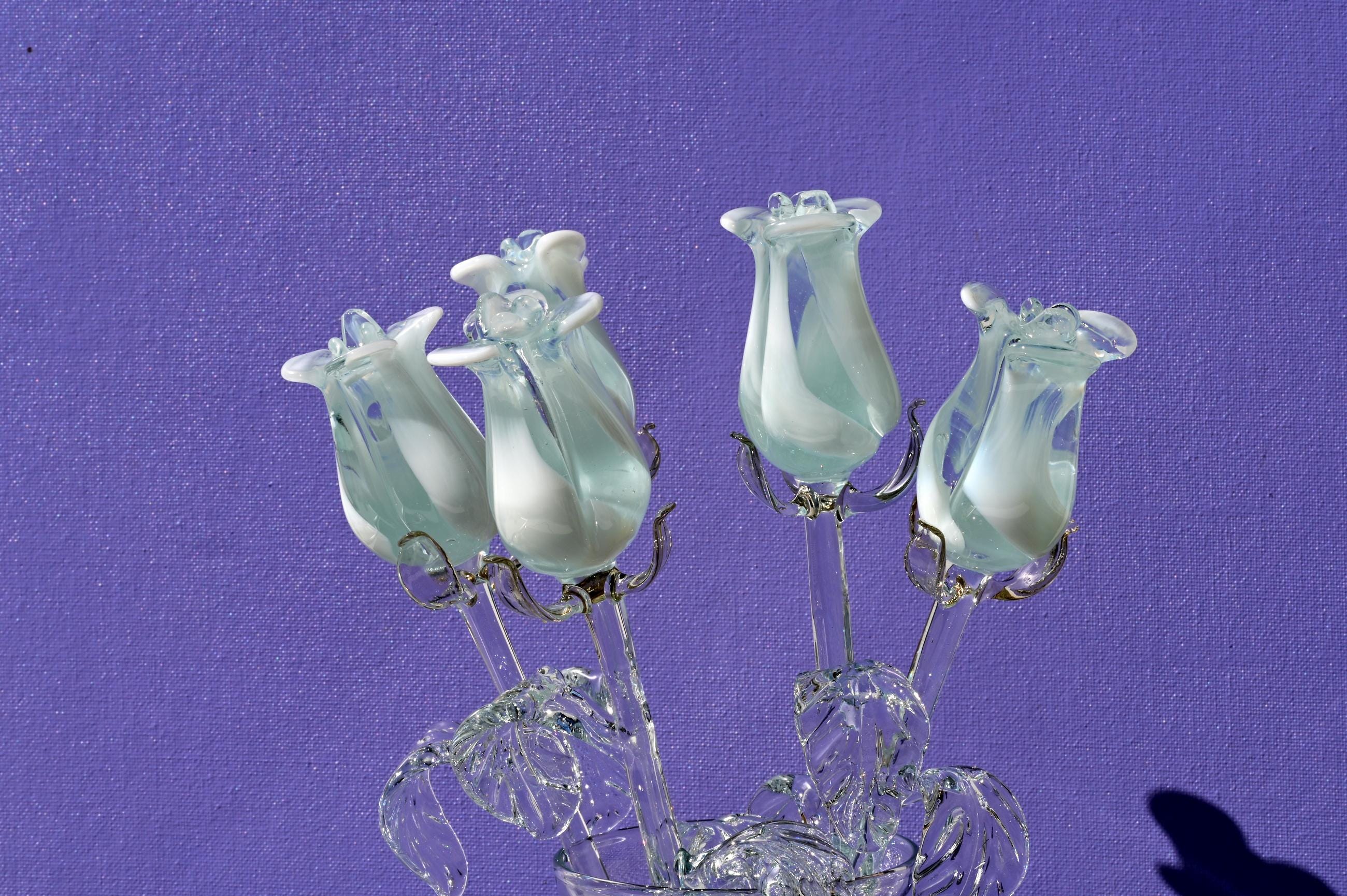 May include: A bouquet of five white glass roses with light blue accents. The roses are arranged in a vase and are on a purple background.