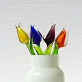 May include: Five glass tulips in a white vase. The tulips are vibrant colors: yellow, blue, purple, and orange. The stems are green glass. The tulips are artfully arranged in a simple, elegant white vase. These handcrafted glass flowers are a unique home decor accent.