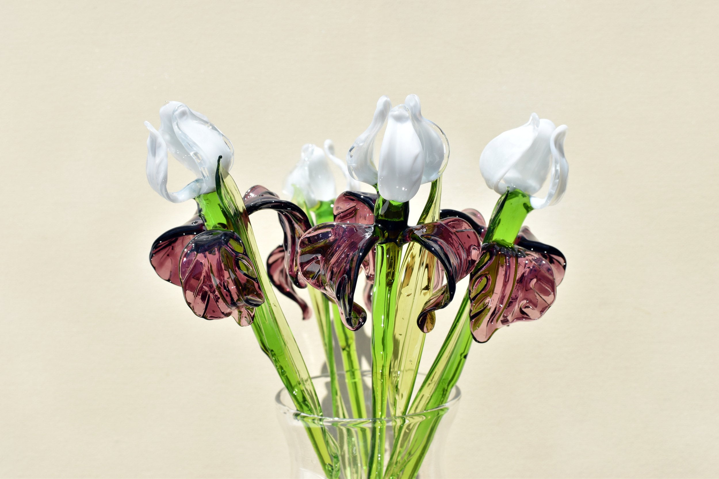 May include: Close-up of a glass art bouquet of irises. Five stemmed irises feature white glass blossoms and deep purple-pink glass leaves. The stems are a mix of light and dark green glass. The bouquet is arranged in a clear glass vase.