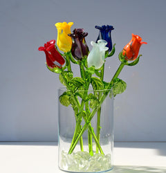 May include: A bouquet of seven glass roses in a clear glass vase with clear glass stones at the bottom. The roses are red, yellow, orange, white, purple, and blue. The stems are green.
