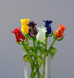 May include: A bouquet of six glass roses in a clear glass vase. The roses are red, yellow, purple, white, blue, and orange. The roses are on green stems with green leaves.