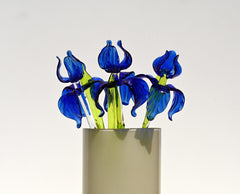 May include: A vase holds three glass flowers. The flowers have blue petals and green stems. The petals are a deep blue with a textured appearance. The stems are a bright green. The vase is a light beige color and cylindrical in shape. The background is a solid white.