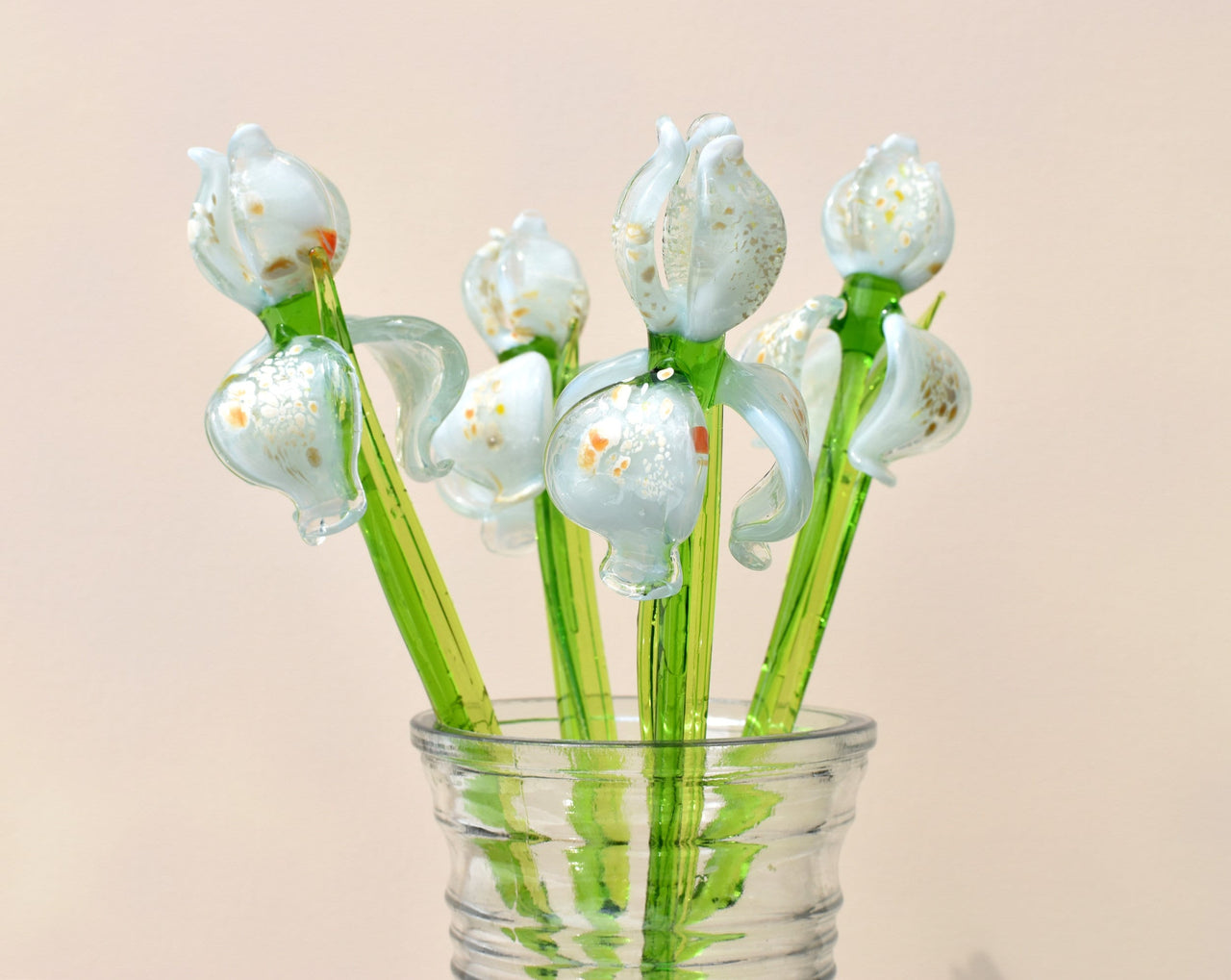 May include: A bouquet of six glass flowers in a clear glass vase. The flowers are white with green stems and have a speckled, iridescent finish. The flowers are arranged in a vase with a textured surface.