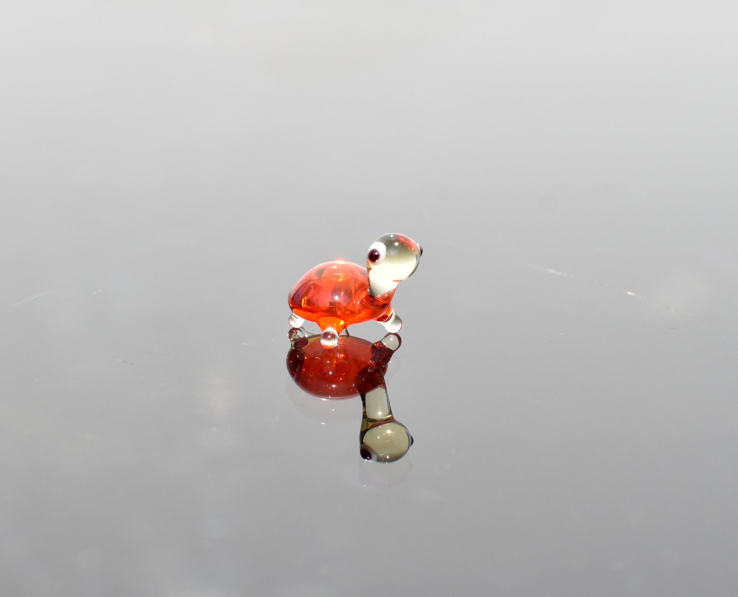 May include: A small glass turtle figurine with a red shell and white eyes. The turtle is reflected in a shiny surface.