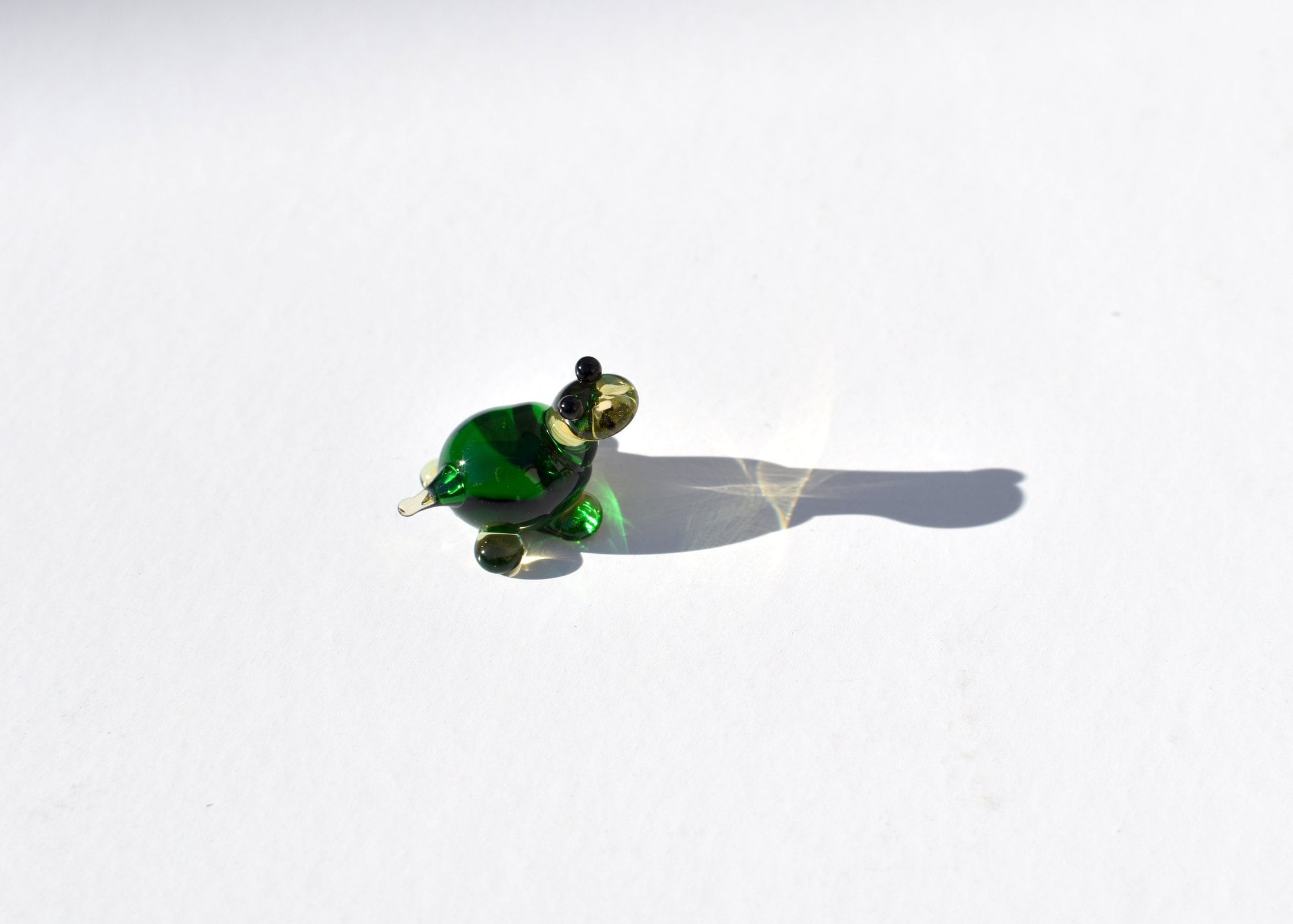 May include: A small green glass turtle figurine with black eyes. The turtle is sitting on a white surface and casting a shadow.