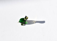 May include: A small green glass turtle figurine with black eyes. The turtle is sitting on a white surface and casting a shadow.