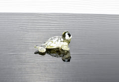 May include: A small glass sea turtle figurine with white and yellow glass. The turtle has black eyes and a white belly. The figurine is sitting on a gray surface.