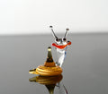 May include: A glass snail figurine with a clear body, orange and red mouth, and black eyes. The snail is sitting on a yellow glass shell.