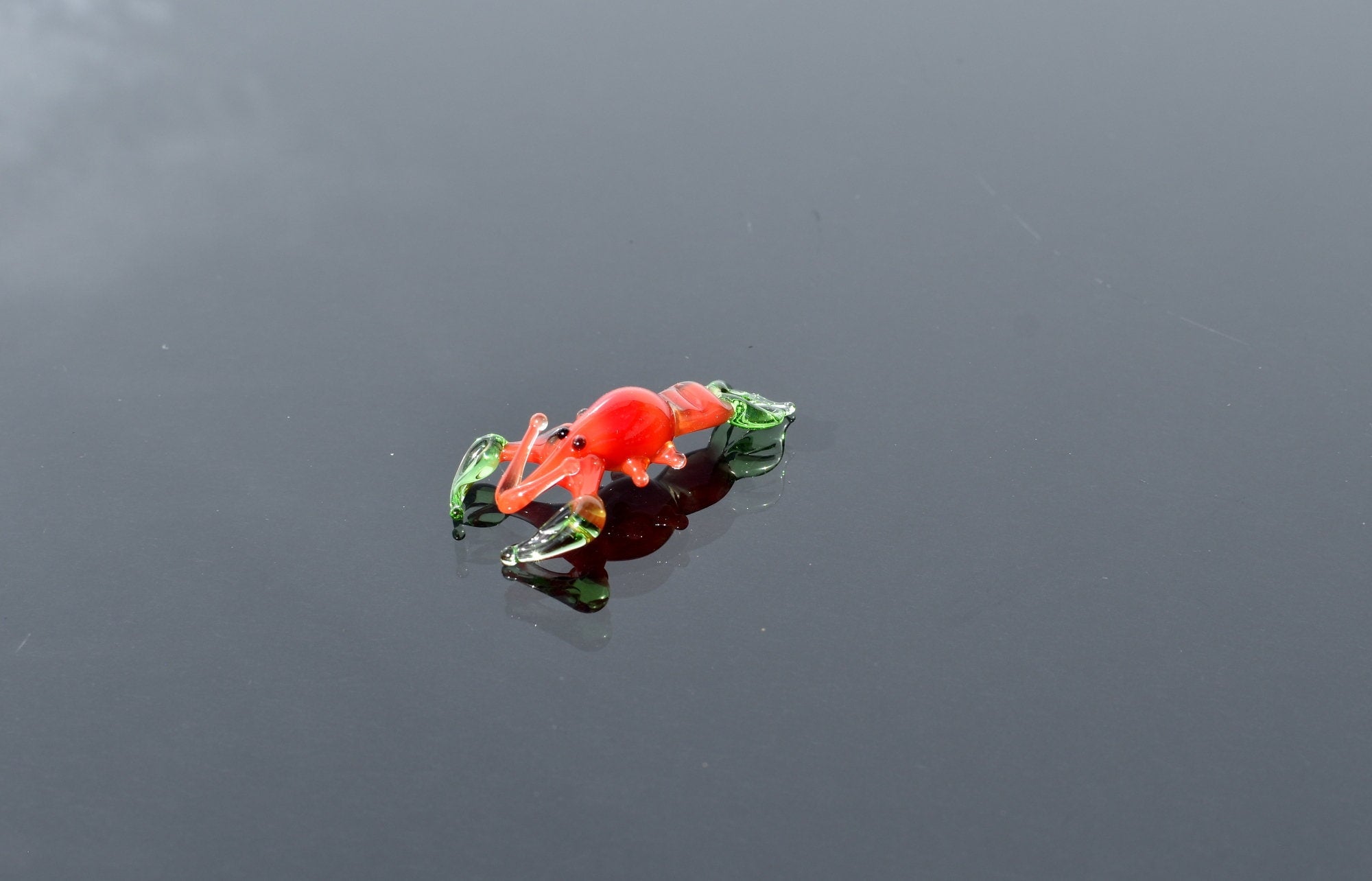 May include: A small glass figurine of a red lobster with green claws. The lobster is posed on its side with its claws outstretched.