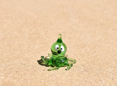May include: A small green glass octopus figurine with white eyes and a black nose. The octopus has eight tentacles and is sitting on a light brown surface.