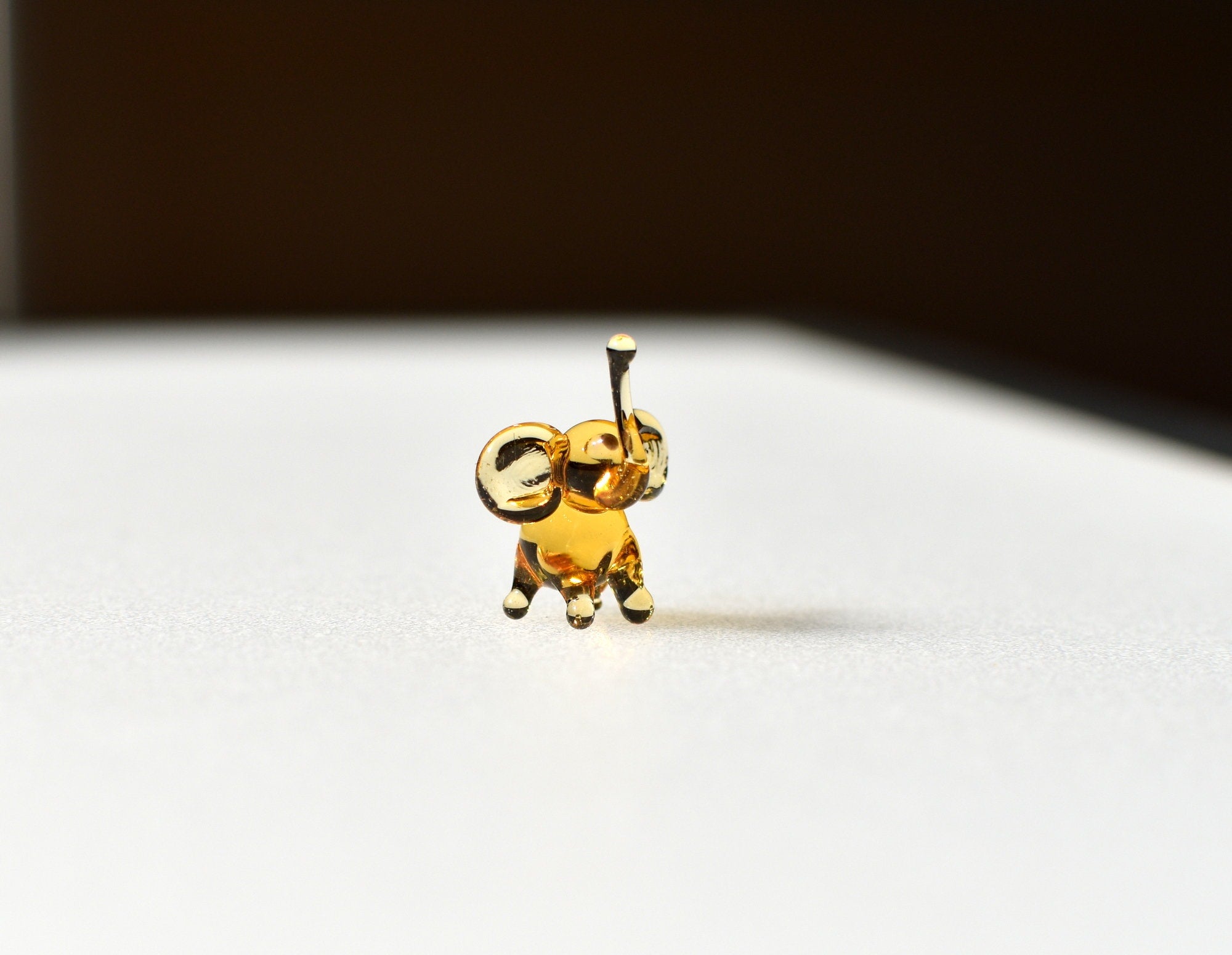 May include: A small, gold-colored glass elephant figurine with a raised trunk. The elephant is standing on a white surface.