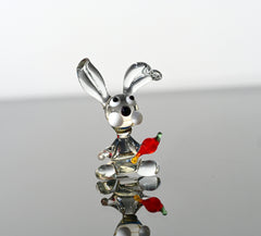May include: A glass rabbit figurine with a carrot in its mouth. The rabbit is clear glass with black eyes and white accents. The carrot is red and orange glass.