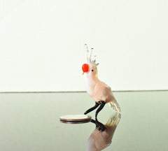 May include: A small glass figurine of a cockatoo with a white body, black legs, and a red beak. The cockatoo is standing on a reflective surface.