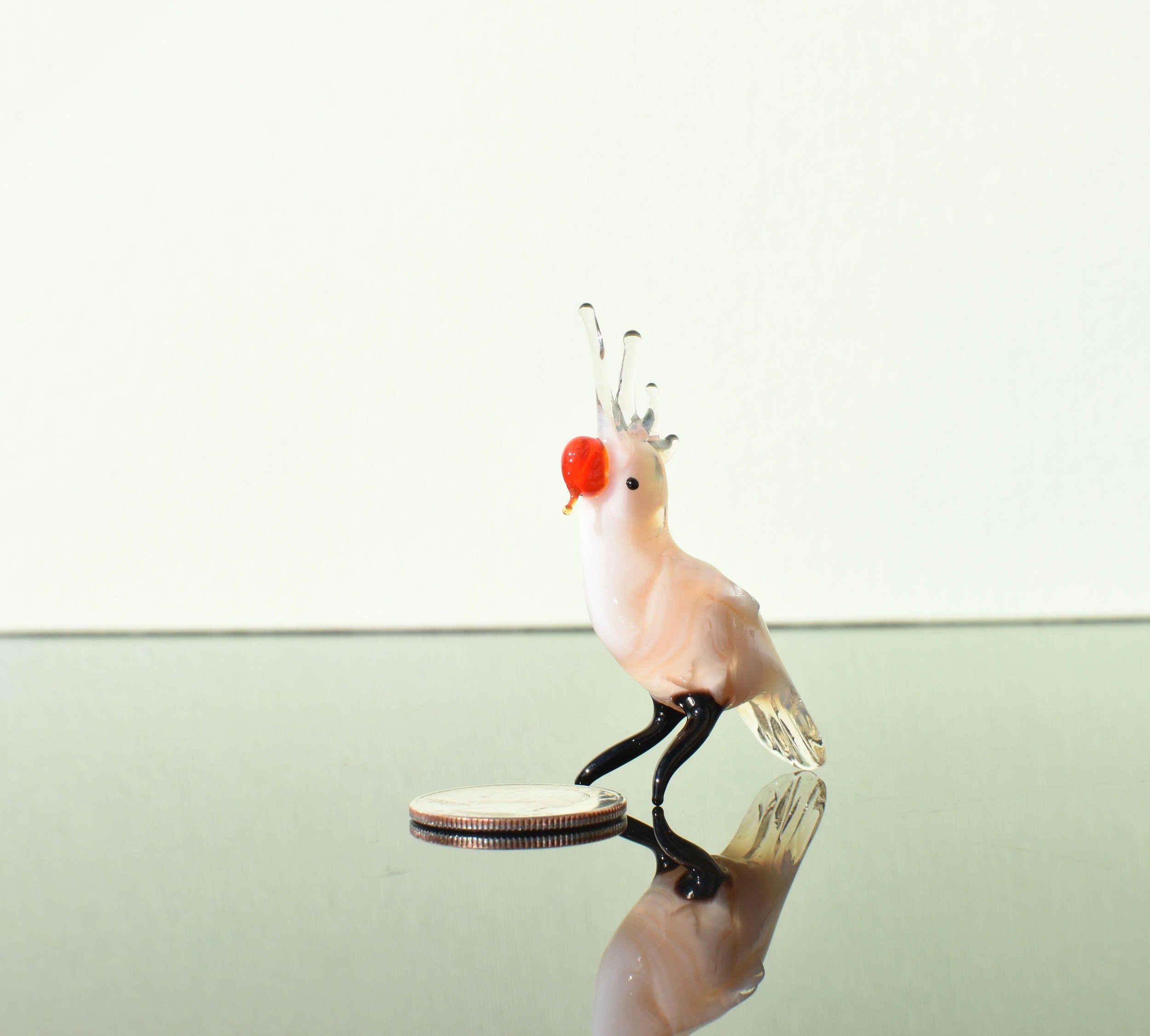May include: A small glass figurine of a cockatoo with a white body, black legs, and a red beak. The cockatoo is standing on a reflective surface.